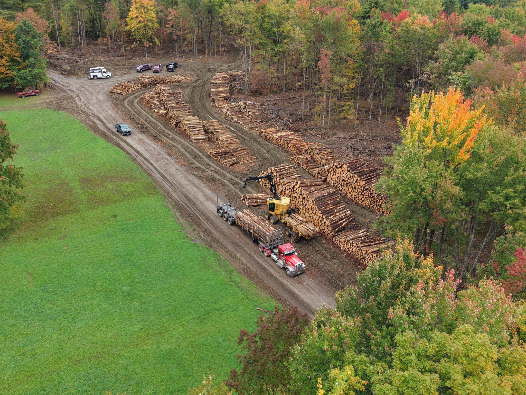 Aerial drone view of a log landing with sorted log decks, a loader, and a loaded log truck surrounded by fall foliage